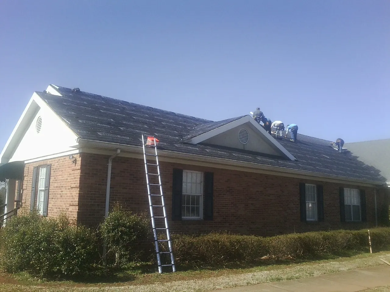 Crew tearing shingles off a brick ranch roof with a ladder set — Metal Roof Repair work by our crew in Evansville
